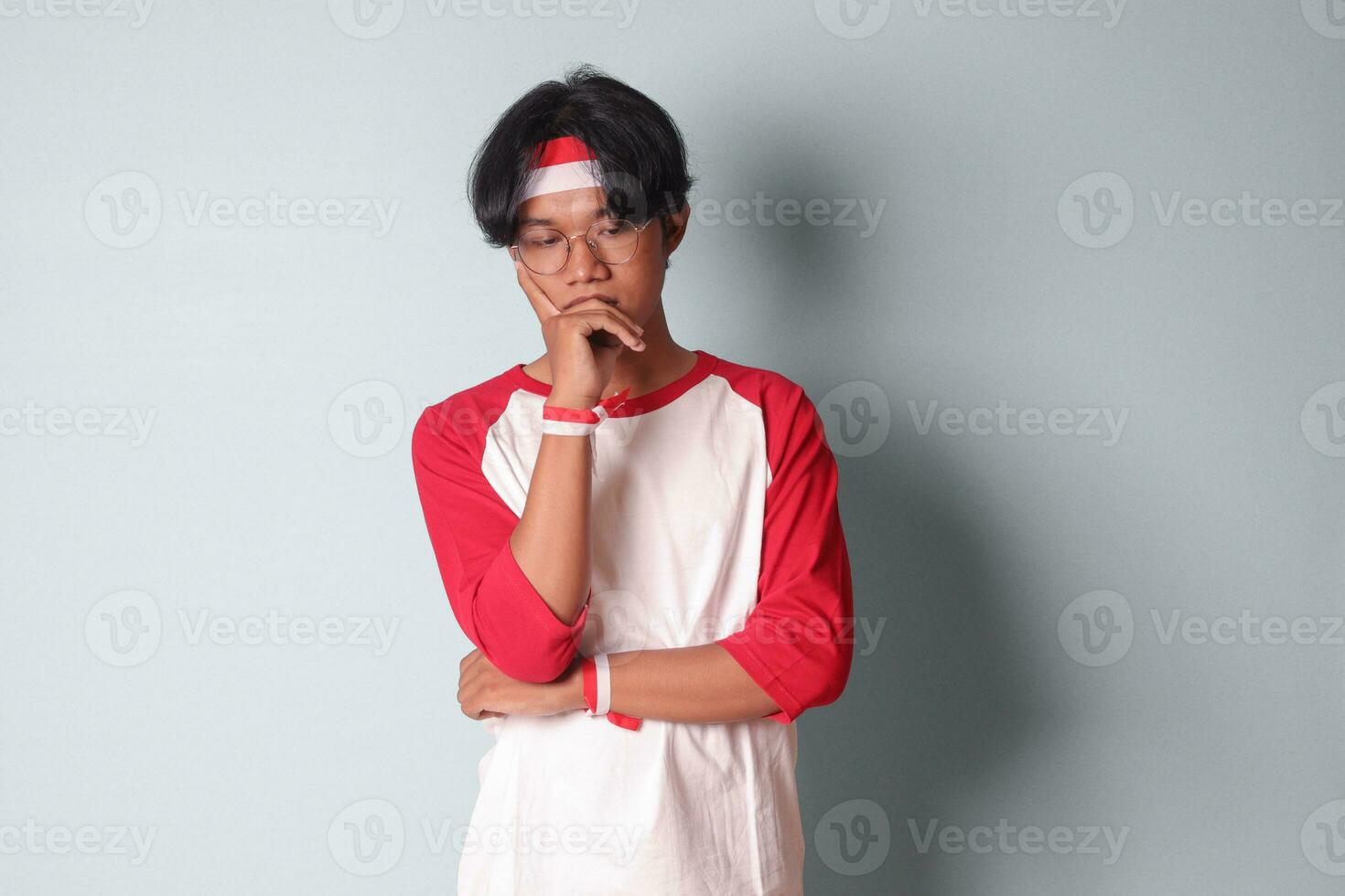 Portrait of attractive Asian man in t-shirt with red and white ribbon on head, standing against gray background, thinking about question with hand on chin photo