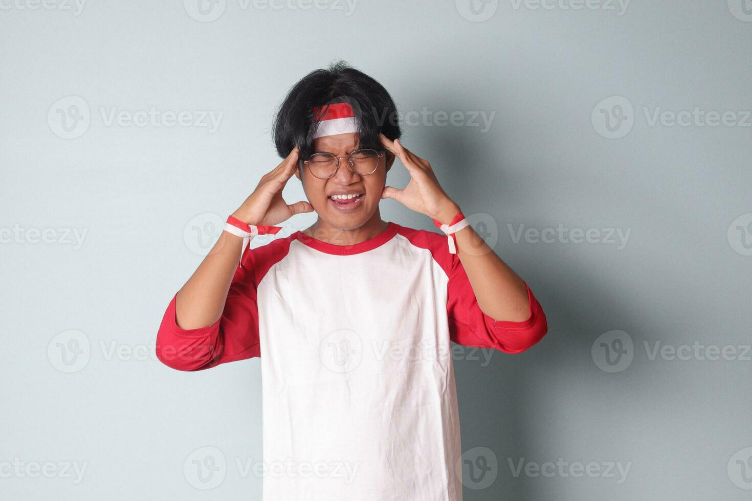 Portrait of attractive Asian man in t-shirt with red and white ribbon on head, screaming with close eyes and wide open mouth, holding hands on forehead. Isolated image on gray background photo
