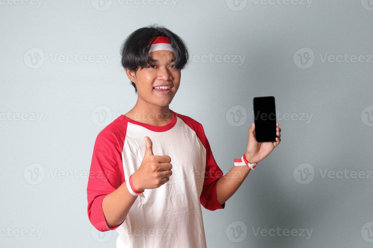 retrato de atractivo asiático hombre en camiseta con rojo y blanco cinta en cabeza, participación y demostración blanco pantalla de móvil teléfono para Bosquejo. aislado imagen en gris antecedentes foto