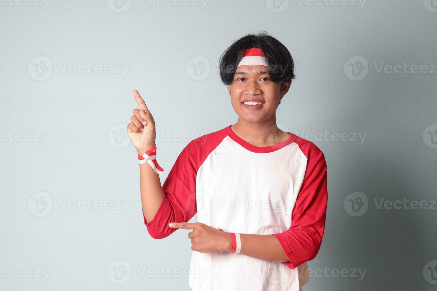 retrato de atractivo asiático hombre en camiseta con rojo y blanco cinta en cabeza, señalando a alguna cosa con dedo. aislado imagen en gris antecedentes foto