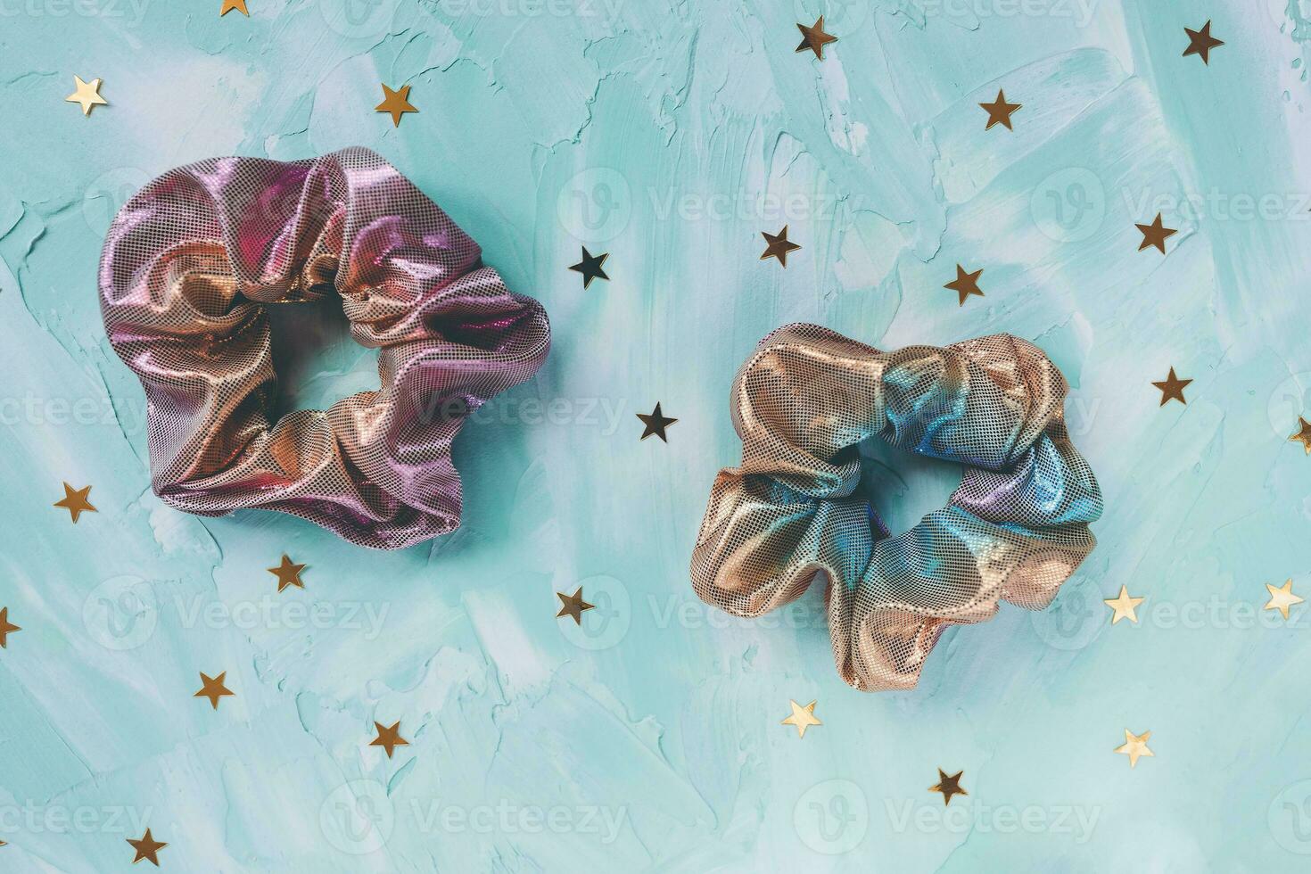 Two trendy holographic iridiscent shiny metallic scrunchies and golden stars confetti on cyan background. Diy accessories for Christmas and New Year holiday party and hairstyles concept, copy space photo