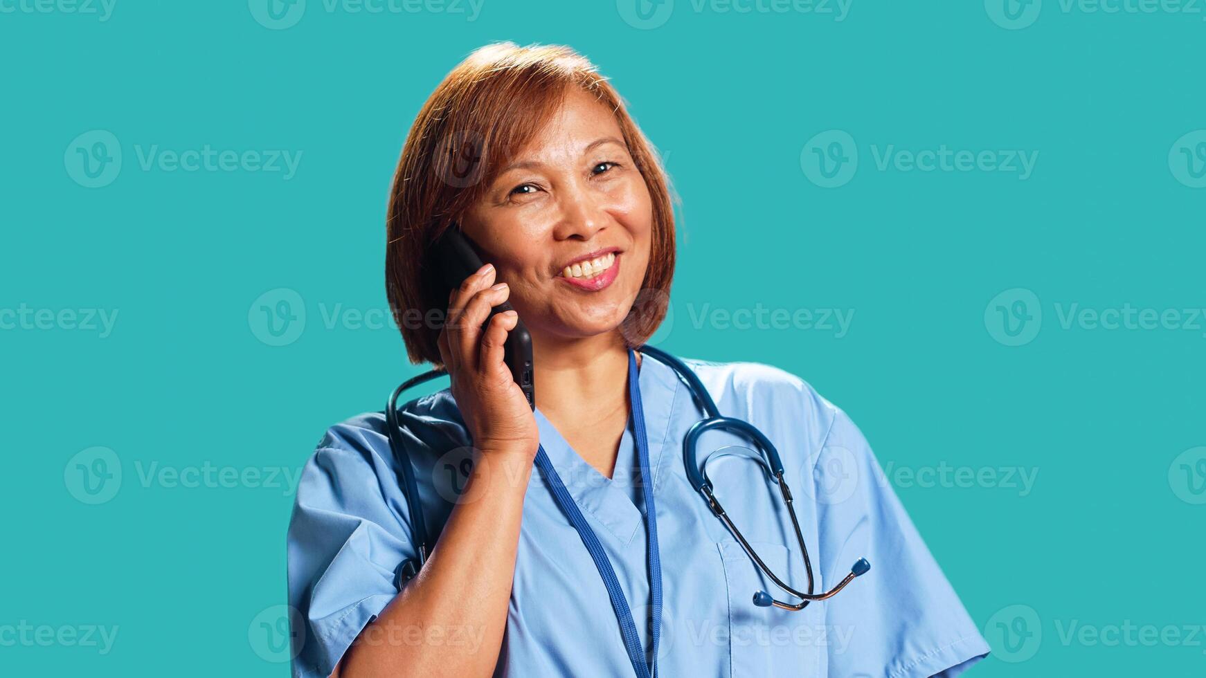 Upbeat BIPOC clinic employee wearing protective uniform having lively chat while on call with friend. Close up shot of nurse happily answering phone while at work, isolated over studio background photo