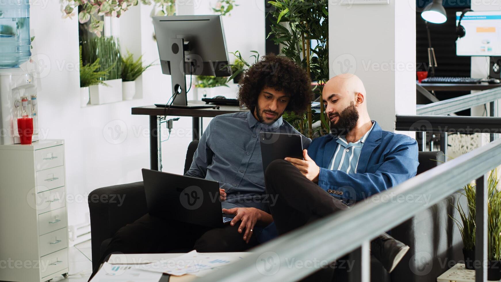 Two employees comparing notes in office, reviewing online company data on laptop and tablet. People planning new presentation for executive board meeting, using official papers. Tripod shot. photo