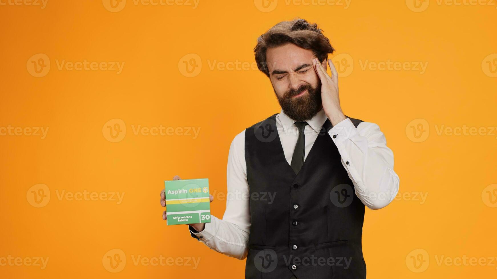 Classy waiter with headache recommends pills on camera, showcasing pack of painkillers to ease pain. Young professional worker feeling unwell and ill, holding medicine treatment box. photo