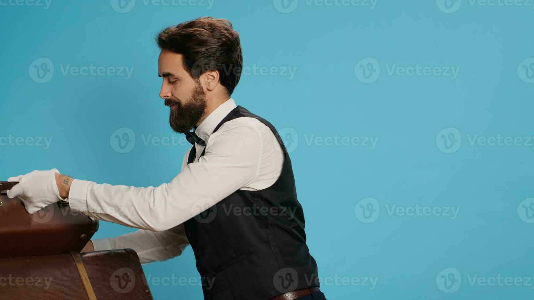 Luxurious employee with briefcase posing against blue background, preparing to carry trolley bags and give assistance to hotel guests. Doorman with formal attire provide classy service. photo