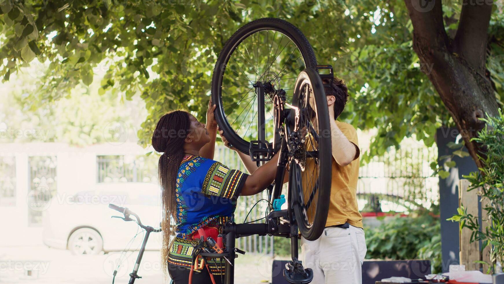 Young enthusiastic caucasian man and african american woman performing maintenance on bike wheels as outdoor summer hobby. Multiethnic couple fastening bicycle tire with professional equipments. photo