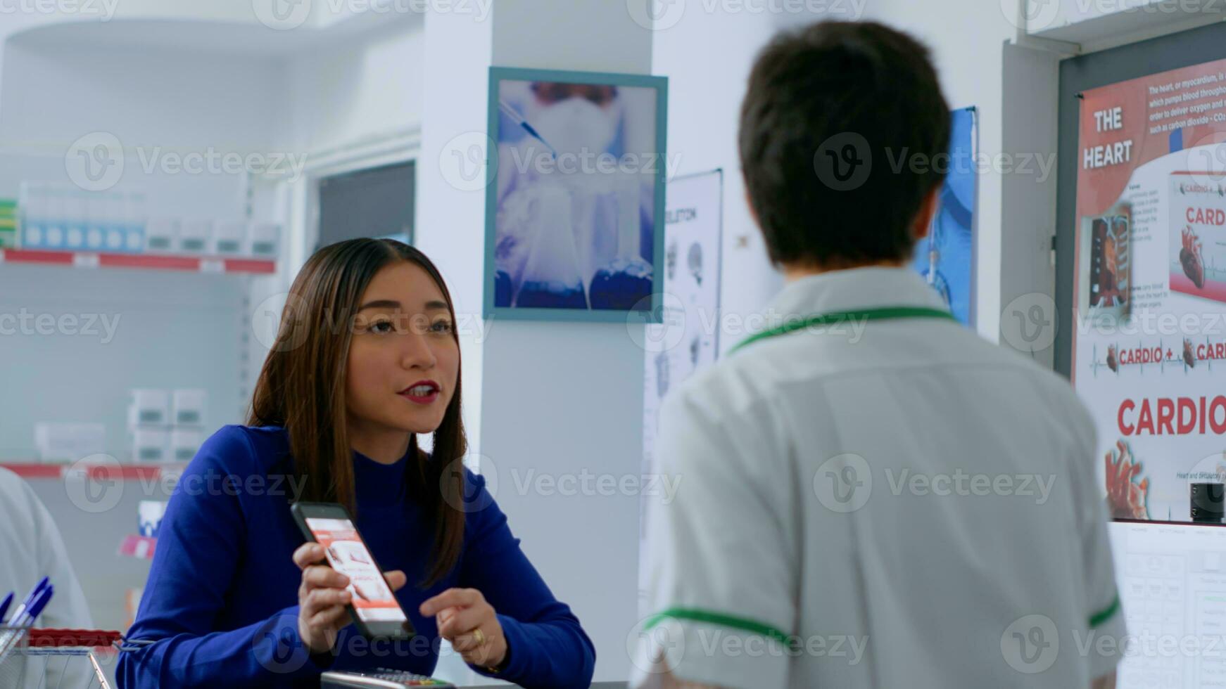 Asian customer at drugstore counter, showing druggist prescribed heart attack prevention medicine and inquiring about required dosage. Wellness expert helping client with needed medicinal product photo