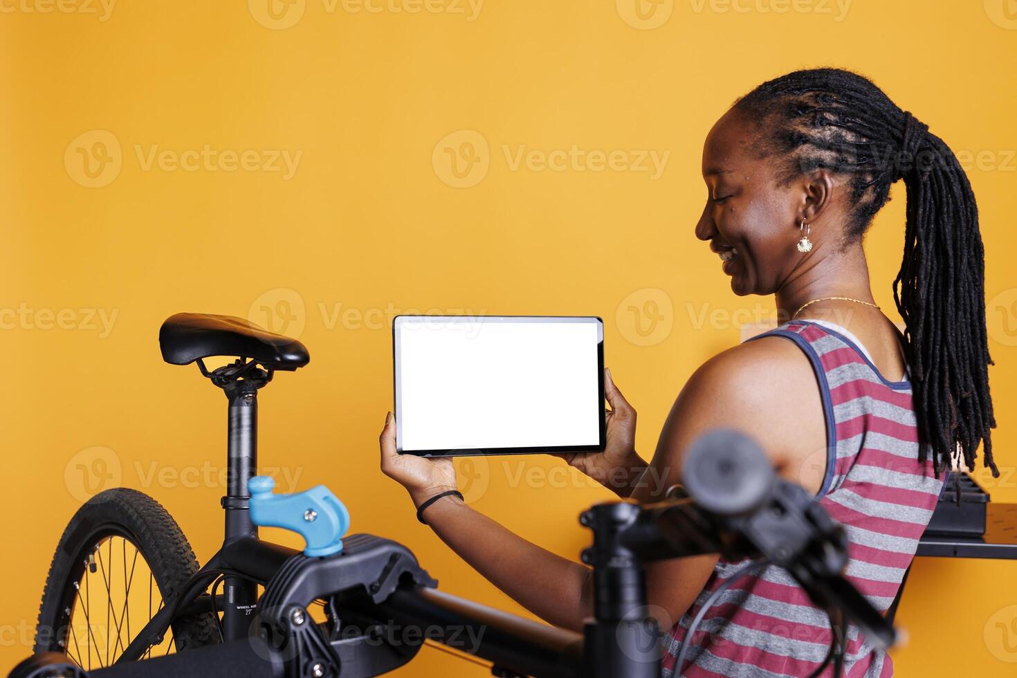 Energetic black woman standing near faulty bicycle and horizontally holding tablet with blank chromakey mockup template. Young female carrying digital device displaying whitescreen. photo