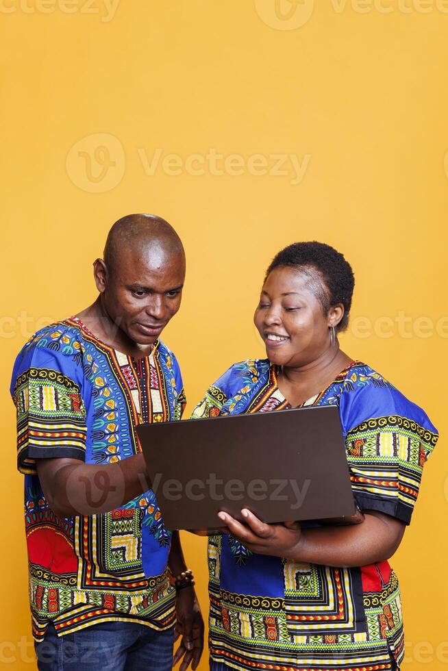 Smiling black man and woman sharing one laptop for work and online pastime activity. Cheerful african american couple wearing ethnic clothes using portable computer in studio photo
