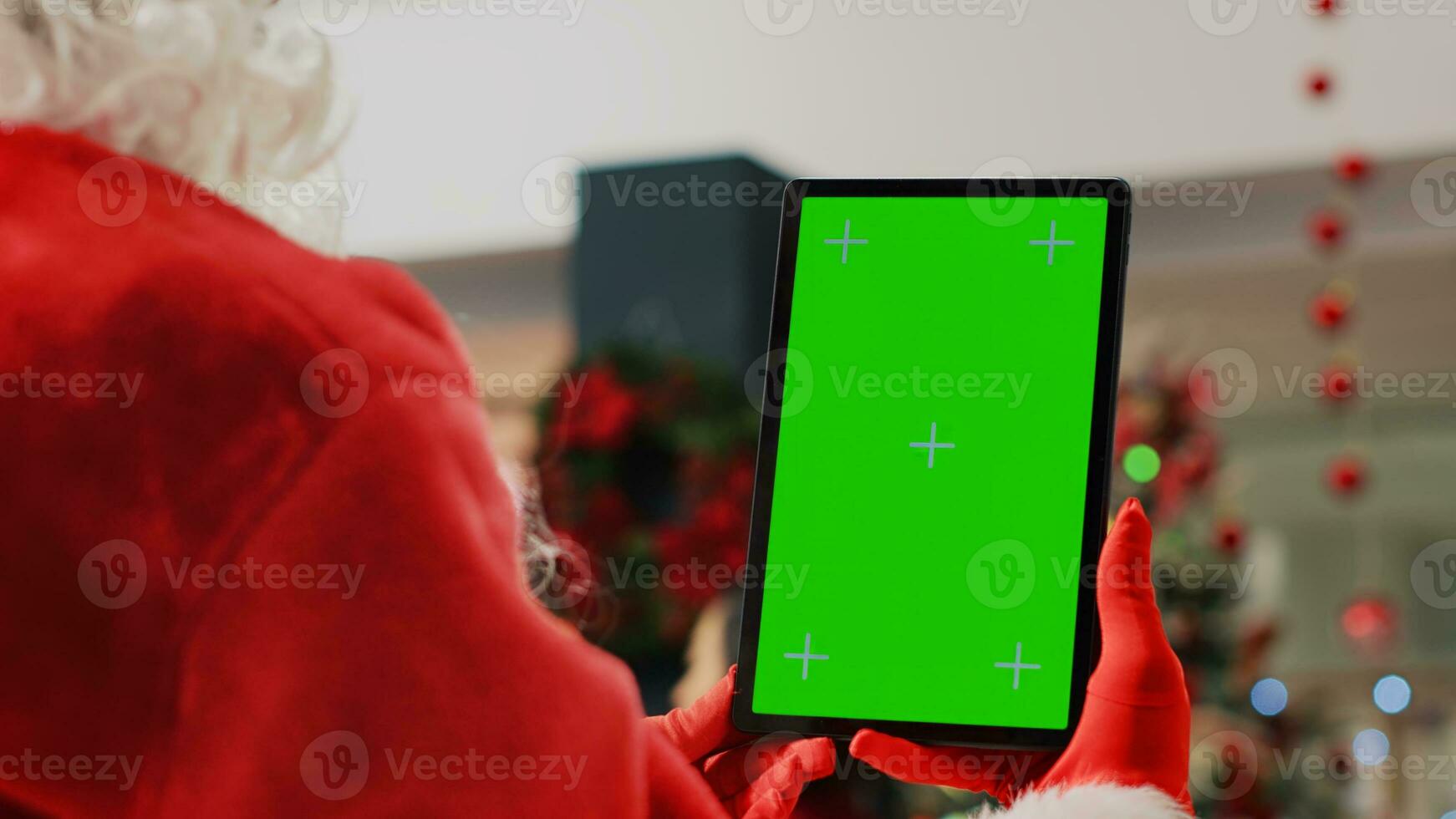 Xmas decorated shopping mall actor impersonating Santa Claus holding chroma key tablet, taking a break from work to watch videos on mockup screen while customers roam around, close up photo