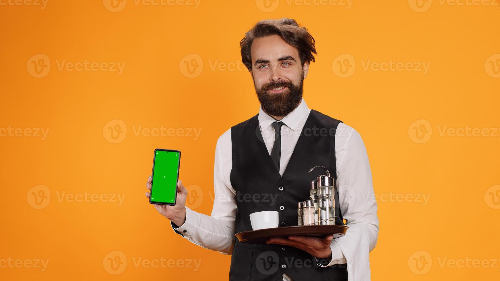 Young waiter shows greenscreen on camera, holding smartphone with blank copyspace display. Friendly elegant fine dining employee indicates isolated empty screen template in studio. photo