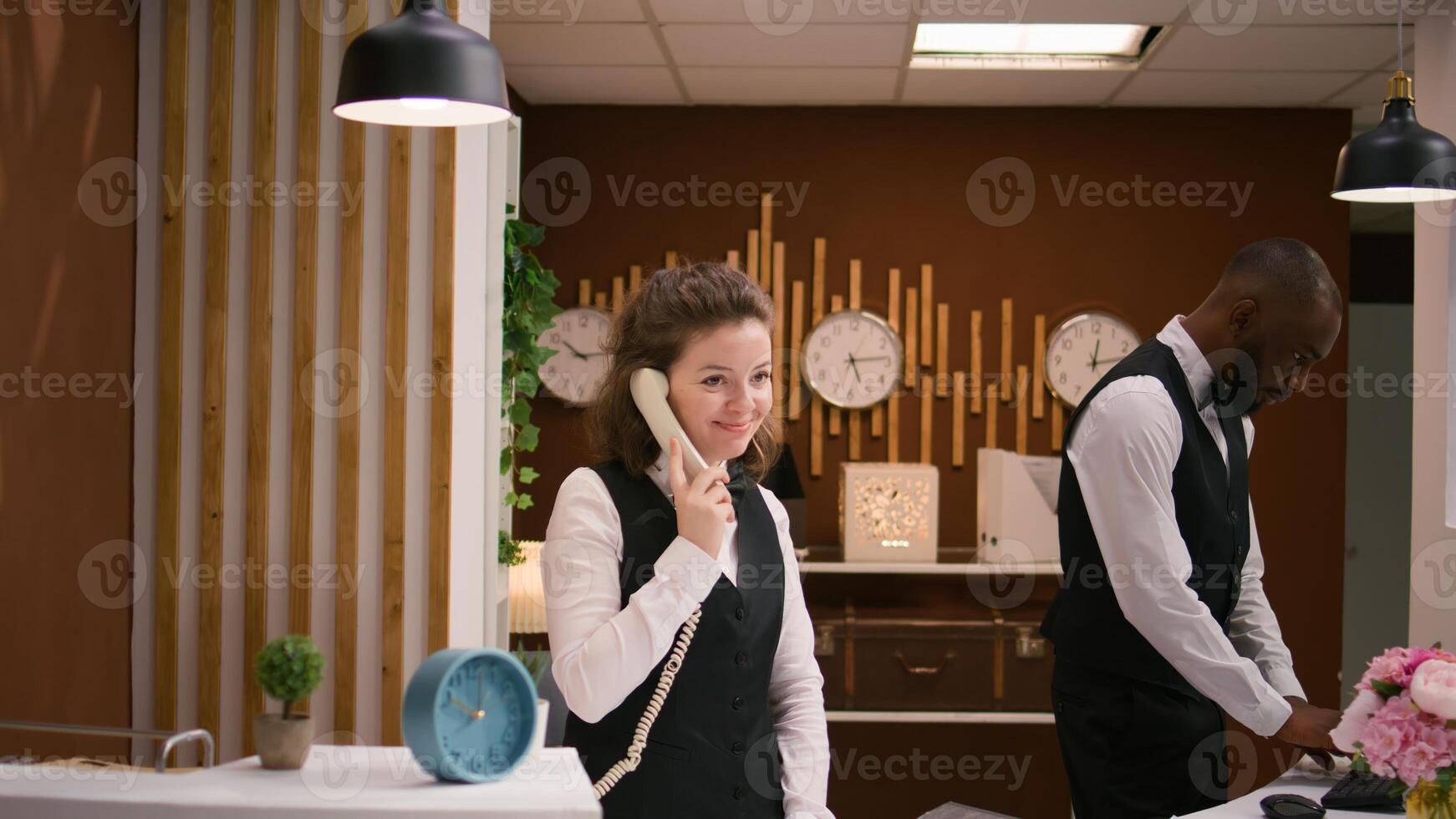 Hotel staff answers calls at reception, using landline phone to make remote bookings at front desk. Friendly receptionist keeping evidence of accommodations and room reservations. photo