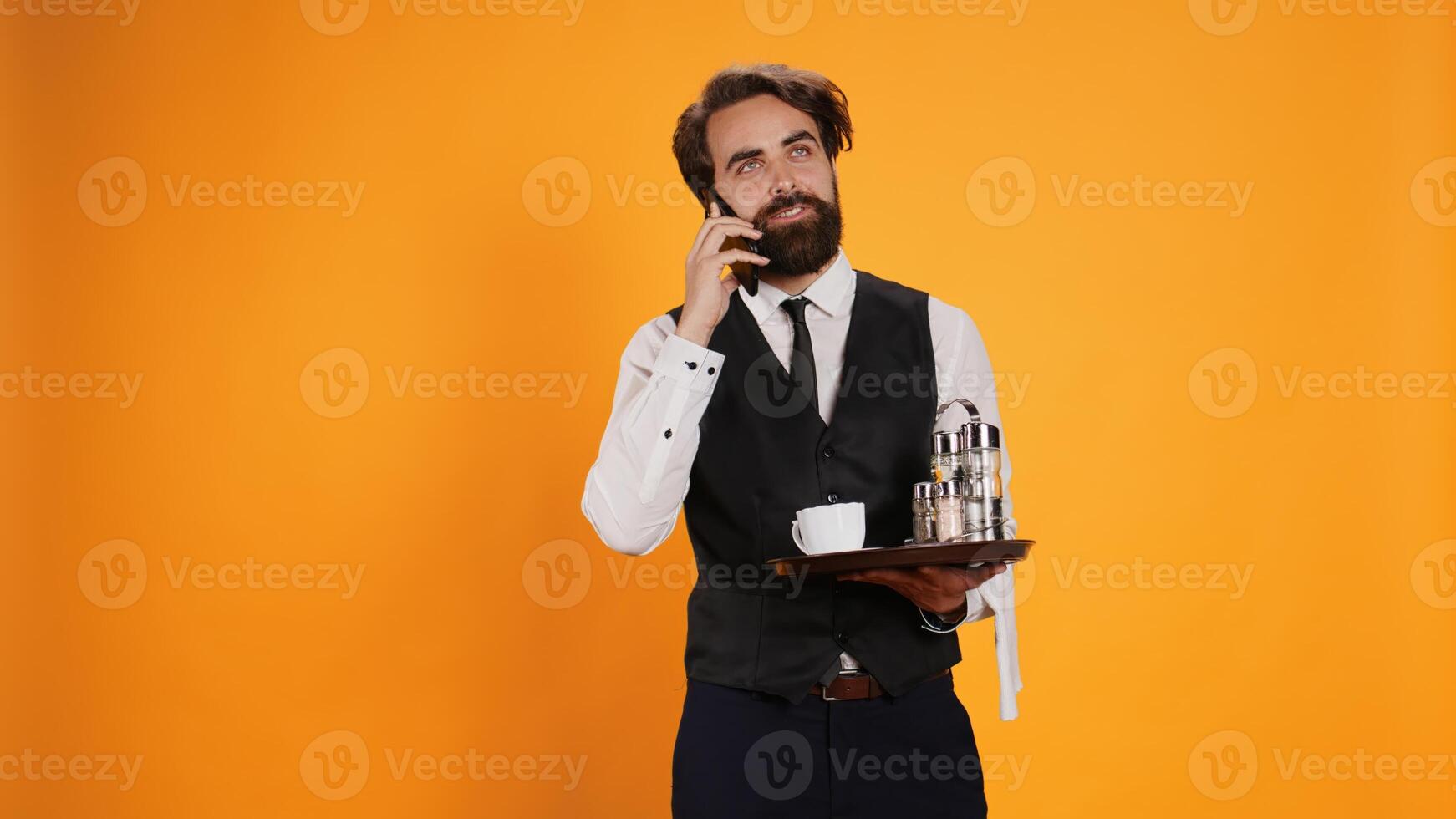 Restaurant waiter on smartphone call making reservations for fancy dinner, working in catering sector against yellow background. Man in formal attire talking to clients on remote phone line. photo