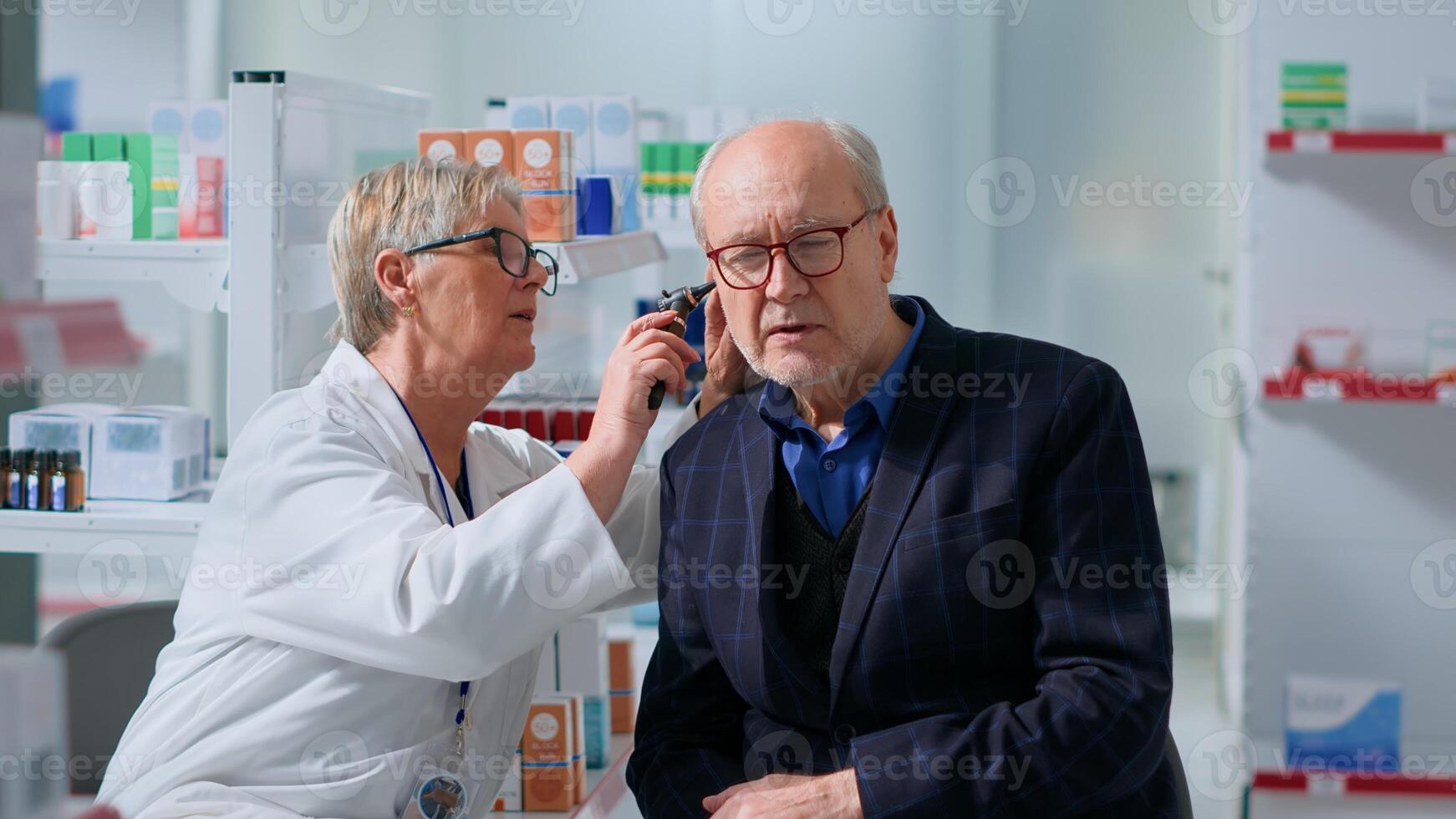 Older pharmacologist in chemist shop doing annual checkup on aged patient, using otoscope to examine ear cannal in professional consultation, preventing hearing damage, excess wax and other issues photo