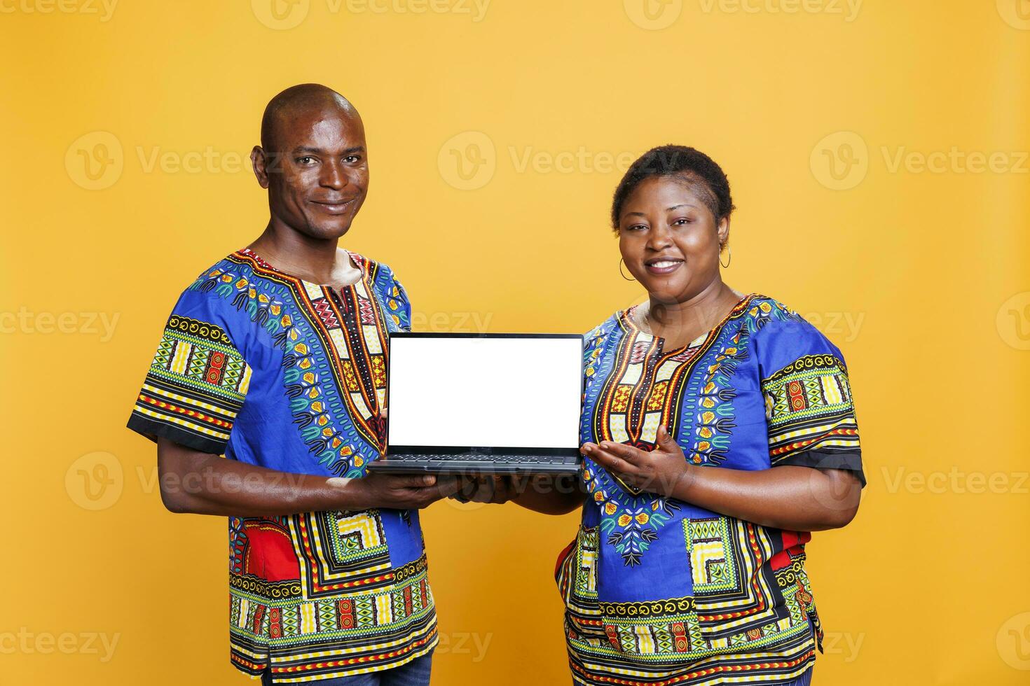 Smiling african american couple wearing ethnic clothes holding laptop with white empty screen mockup. Man and woman showcasing portable computer with blank display and looking at camera photo