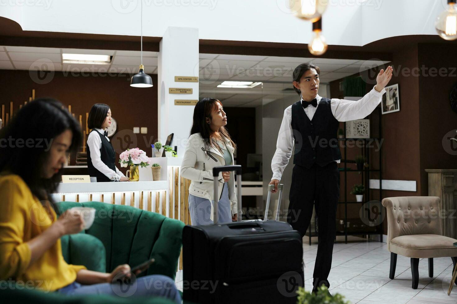 Asian male receptionist giving tourist directions to room in hallway of lavish resort. Woman sitting in hotel lounge area drinking coffee, waiting to make room reservation photo