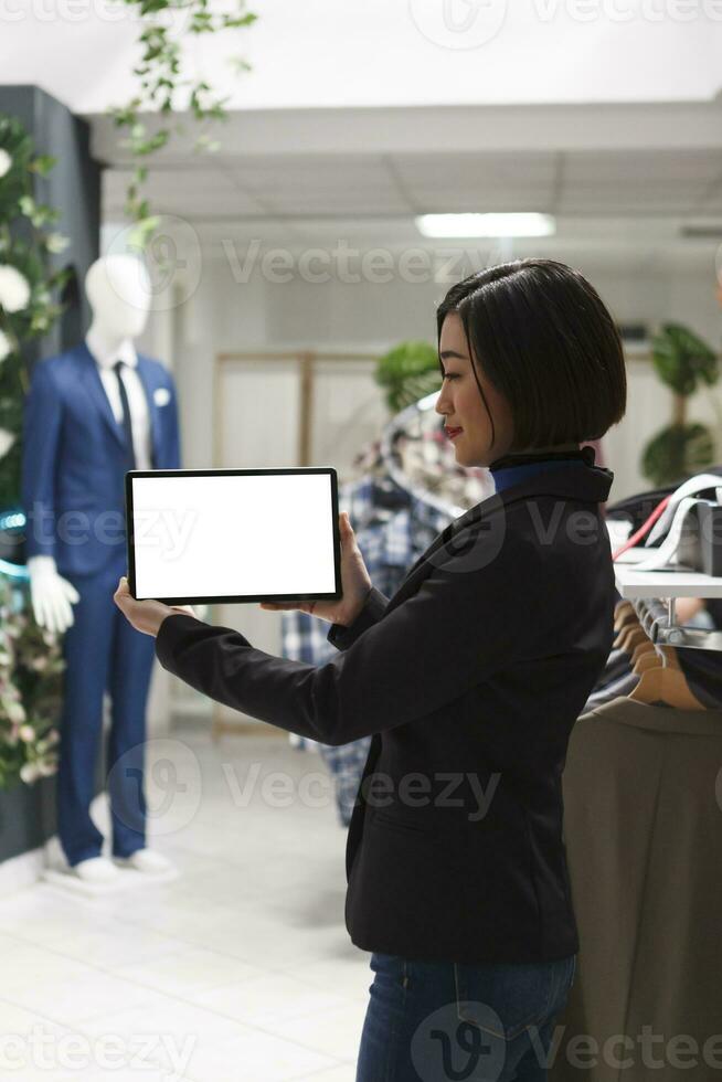 Shopping center boutique asian woman seller holding digital tablet with blank screen, offering assistance. Clothing store young consultant showcasing portable gadget with empty display photo