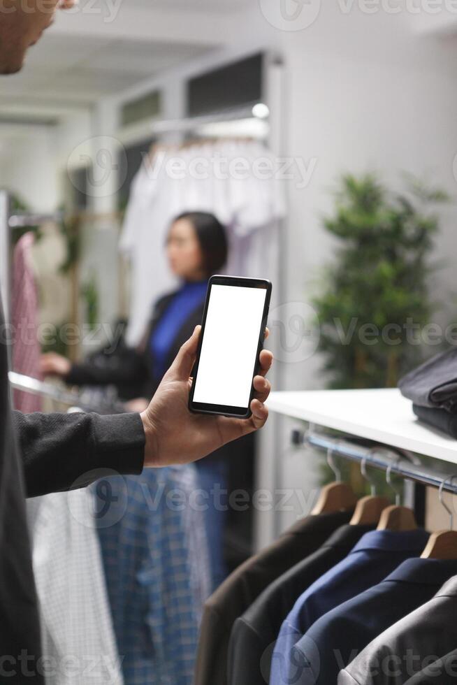 Clothing store customer man hand holding smartphone with blank white screen mockup, checking apparel on boutique website. Shopping mall client using mobile phone with empty touchscreen photo