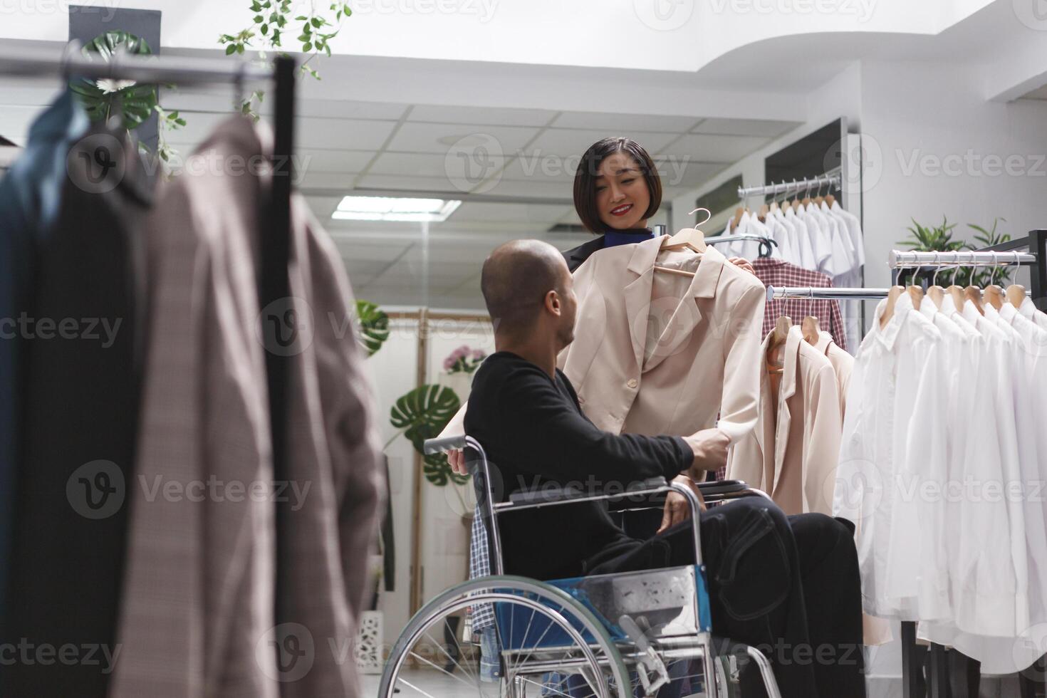 Clothing store smiling asian woman consultant helping arab man with disability in exploring formal womenswear rack. Seller assisting buyer in wheelchair in selecting beige jacket photo