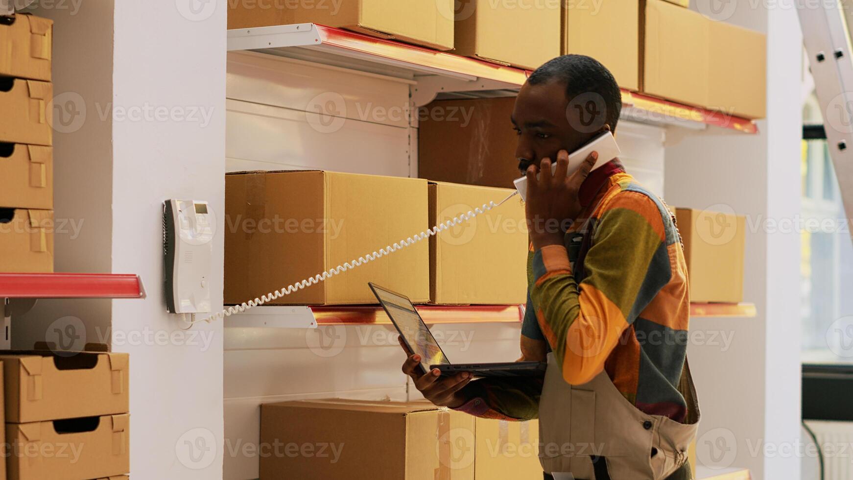 Supervisor using landlinephone call, discussing about supply chain and production with goods. Young adult verifying products before doing shipment logistics, storage room. Handheld shot. photo