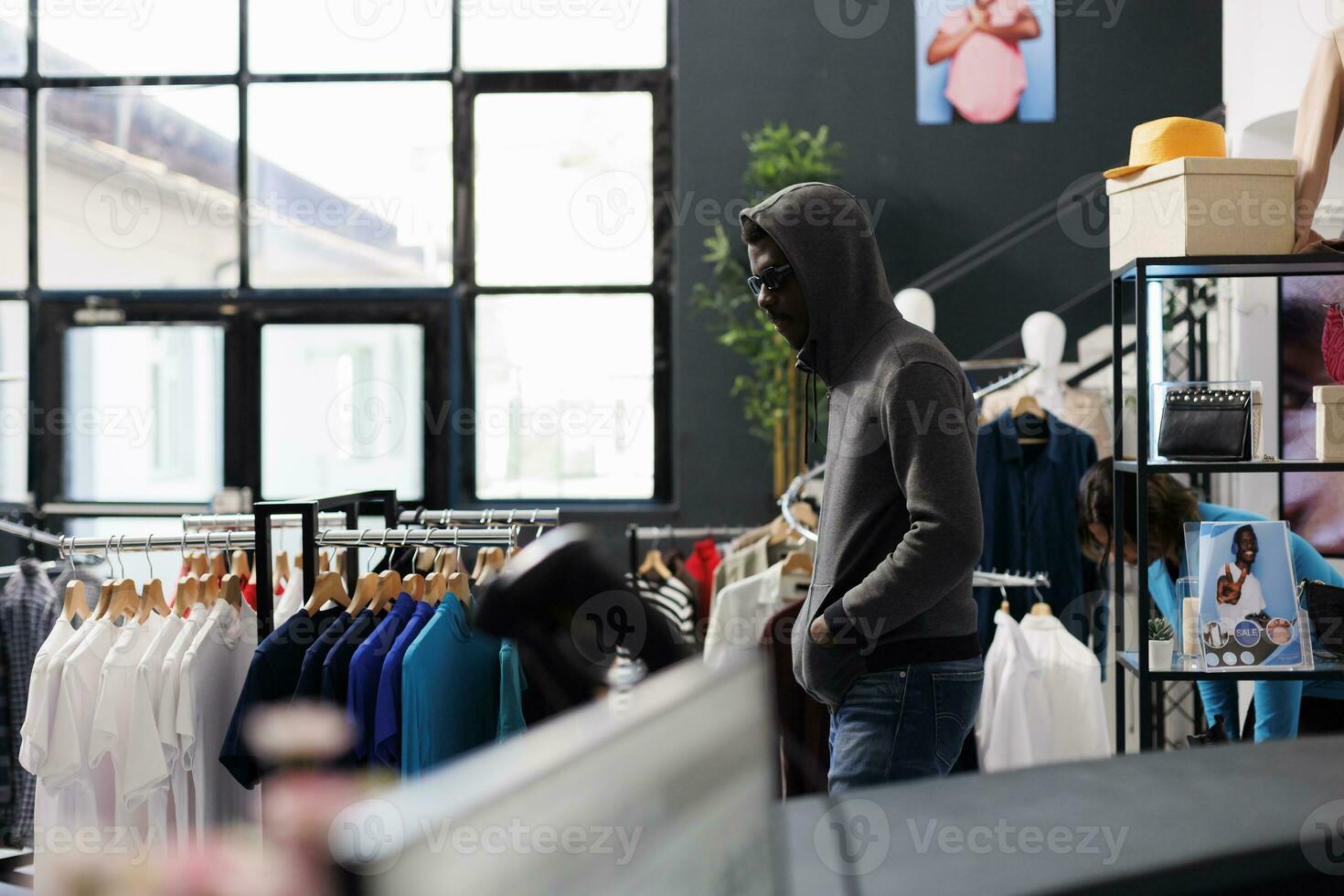 africano americano hombre vistiendo capucha y lentes molesto a robar de moda ropa desde moderno boutique. ladrón mirando alrededor a ver Si alguien acecho él mientras robando mercancías foto
