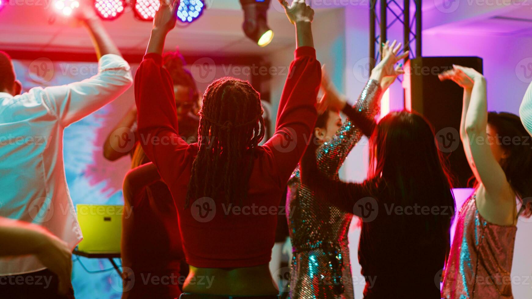 Funky persons dancing at nightclub, cheering for DJ and electronic music mix on stage. Group of people partying and jumping on dance floor with discolights and audio panel. Tripod shot. photo