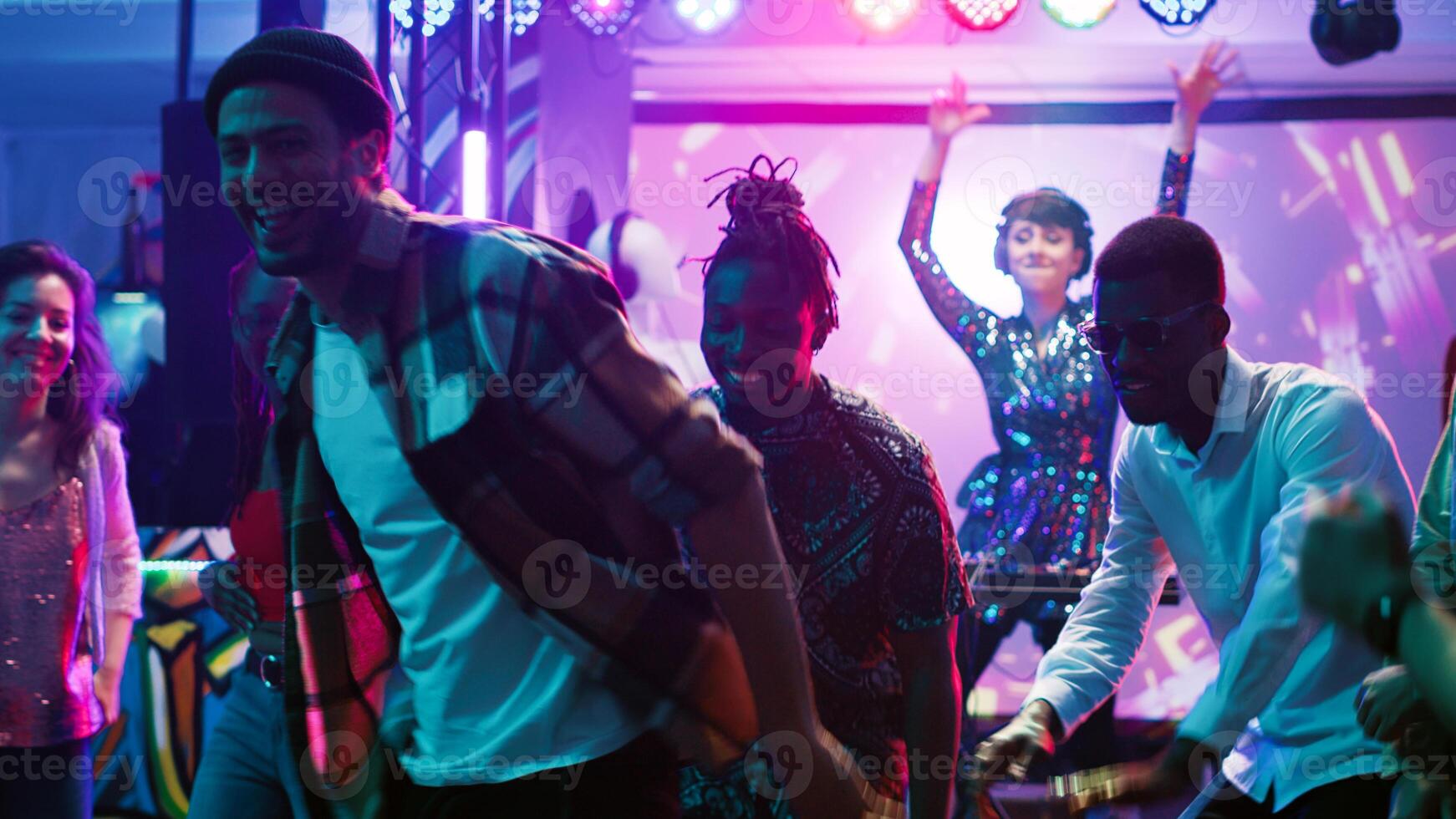 Diverse people partying at club, enjoying live DJ music on bright colorful dance floor. Group of friends having fun at party with stage lights and electronic sound, dancing at nightclub. Tripod shot. photo