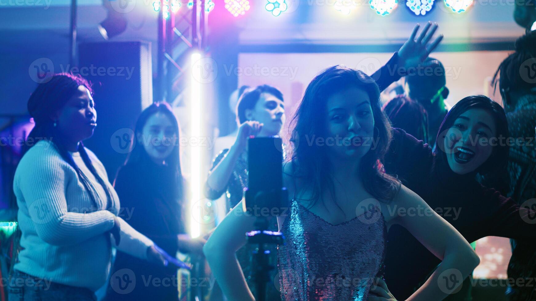 Young adult filming video at party, using mobile phone to record vlog of group of people having fun at club. Smiling person enjoying night out partying with friends, entertainment. photo