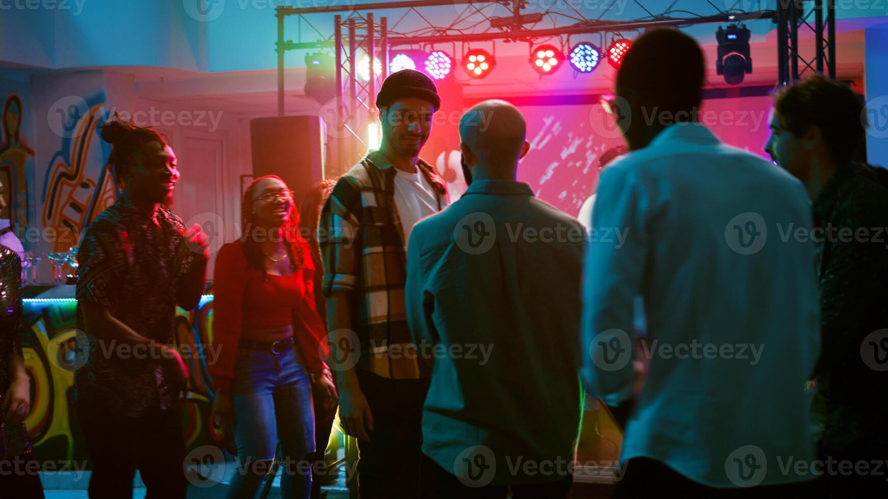 Happy adults improvise breakdance battle at club party, jumping around on dance floor with disco music and spotlights. Group of people showing off cool moves at nightclub. Handheld shot. photo