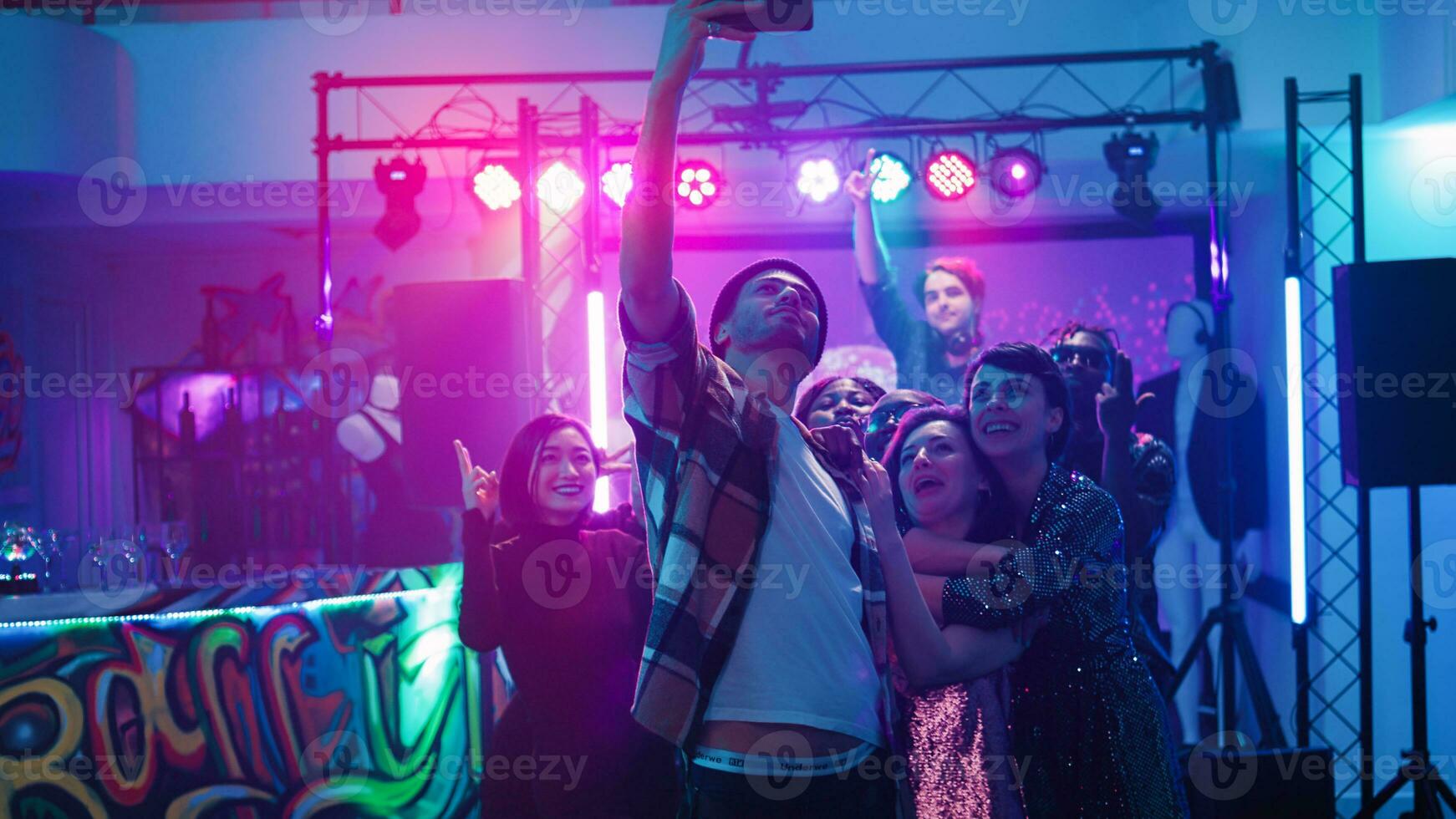 diverso grupo de adultos tomando fotos en danza piso debajo vistoso focos, teniendo divertido a fiesta celebracion. alegre hombres y mujer disfrutando disco evento rodaje vídeos Mano disparo.