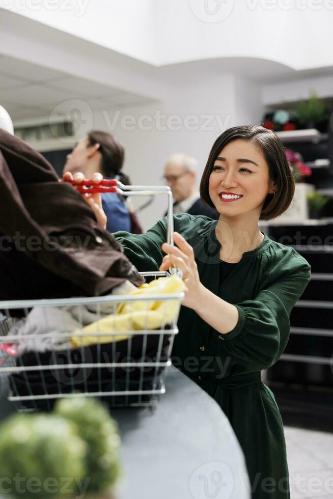 contento sonriente joven hembra cliente en pie en ropa Tienda dando a cajero compras cesta lleno de ropa. alegre adicto a las compras mujer disfrutando compras en Moda boutique. Al por menor terapia foto