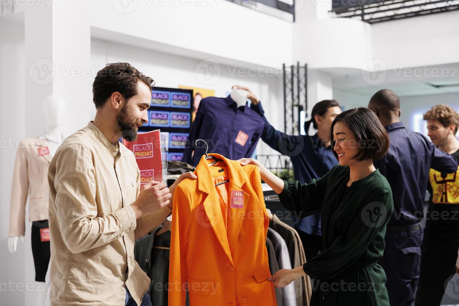 Happy diverse couple shopping on Black Friday in clothing store, looking at orange formal jacket with red sale tag and discussing price. Husband helping wife to choose clothes during seasonal sales photo