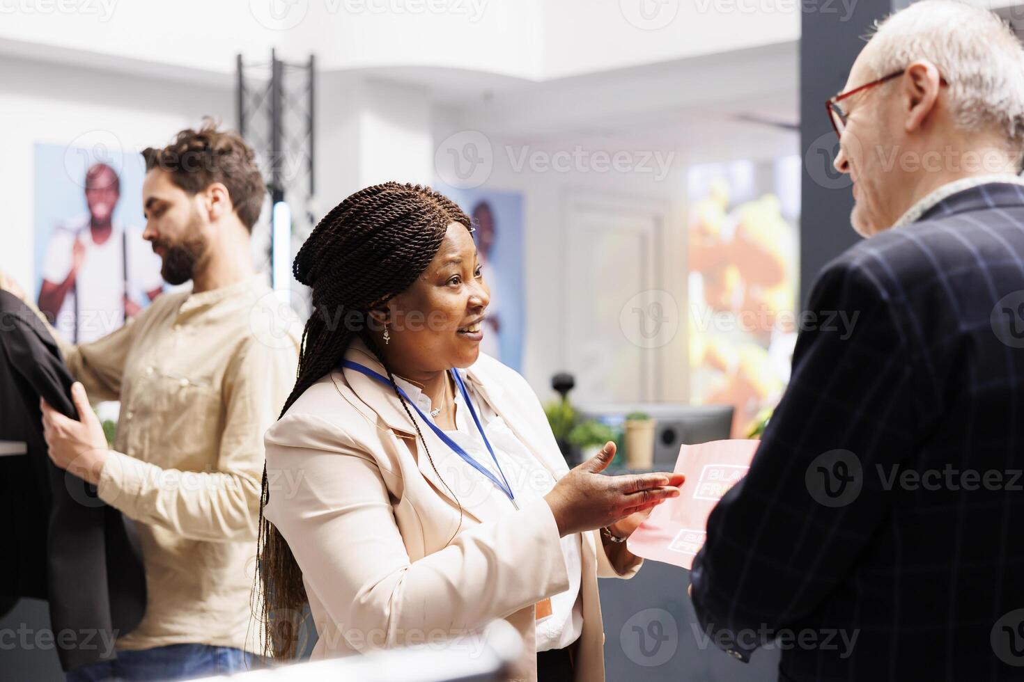 Friendly smiling African American woman shop sales assistant explaining senior man customer Black Friday shopping rules in clothing store. Retail worker holding sales flyer talking with shopper photo