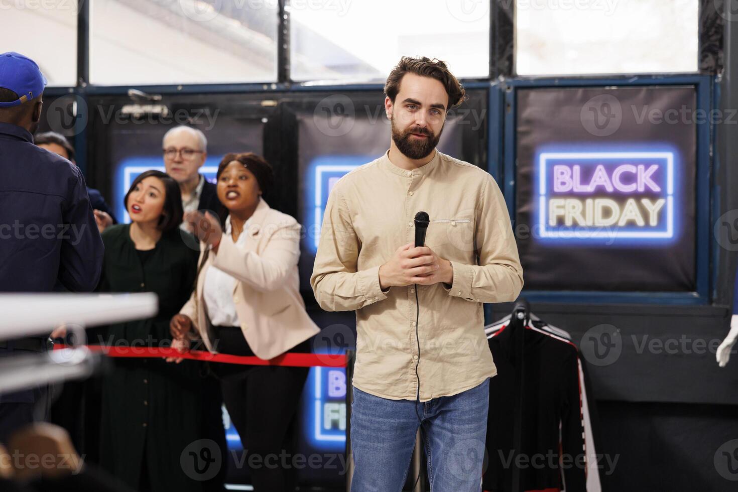 Young man reporter making TV reportage at local shopping mall during Black Friday sales, talking into microphone reporting on shopping madness in stores and how people behaving during seasonal sales photo