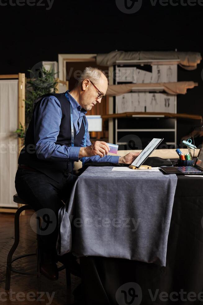 Fashion designer ready to buy sewing materials he needs for upcoming client bespoken comission after finding inspiration online. Elderly suitmaker purchasing tailoring materials with credit card photo