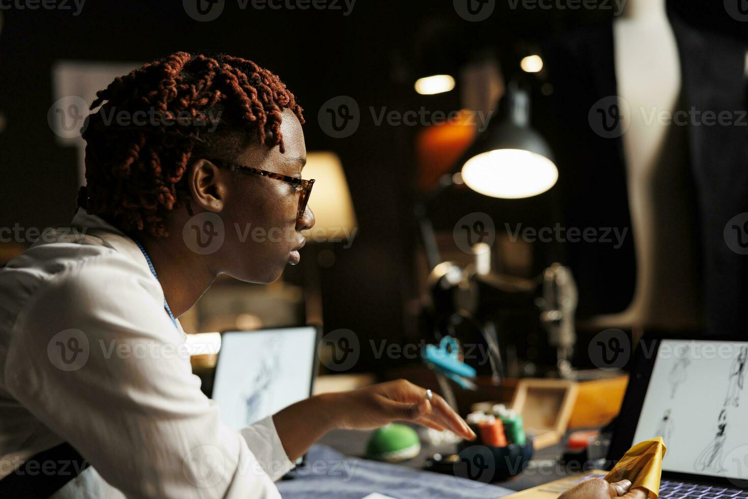 cerca arriba de africano americano modisto vistiendo lentes mirando en línea para Moda diseño inspiraciones. costurera en sastrería investigando creativo conceptos para alta alta costura colección foto