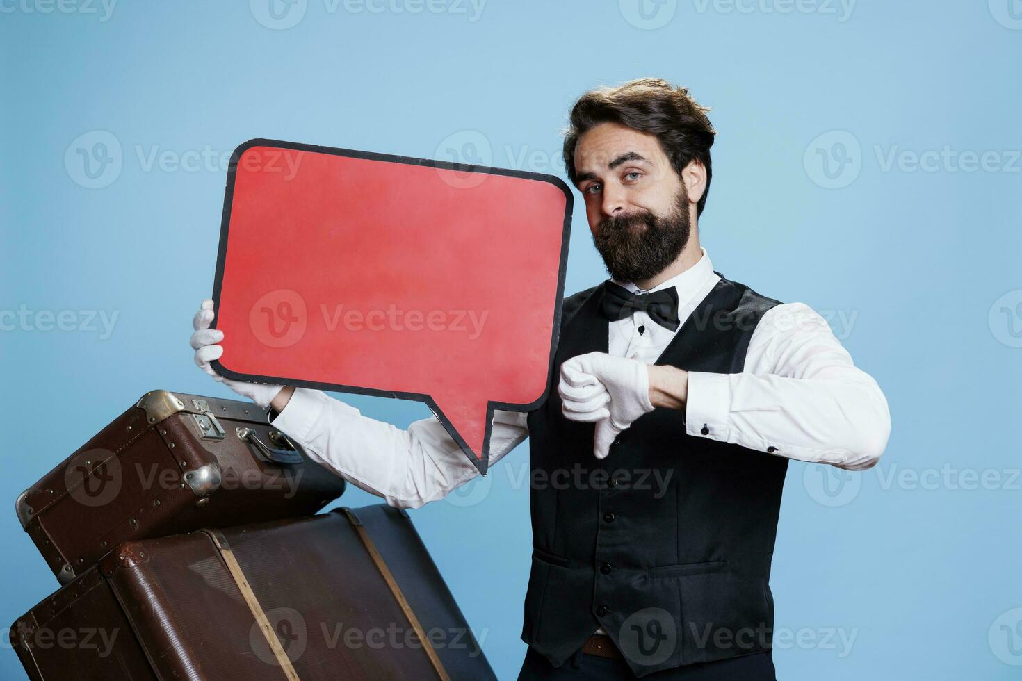 Classy bellboy with speech bubble icon gives thumbs down on camera while he tries to present advertisement on blank billboard. Hotel concierge uses red cardboard sign to express disappointment. photo