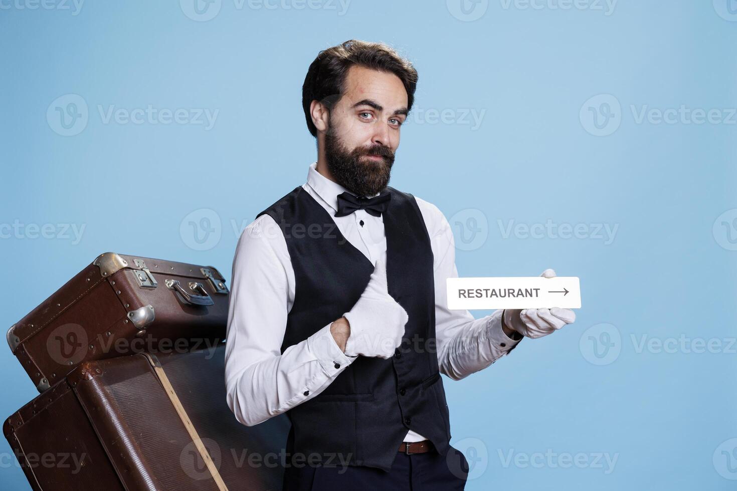 Staff member poses on camera while pointing at restaurant sign to direct visitors. Young sophisticated bellhop presents dining room indicator emblem, wall pointer symbol. photo