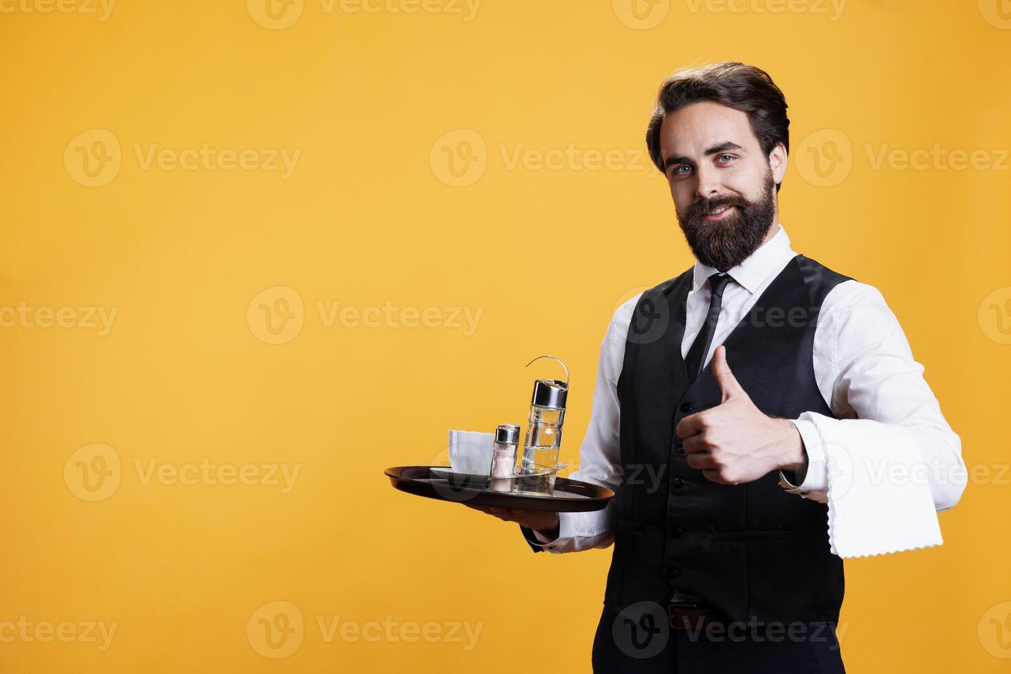 Experienced butler gives thumbs up in studio, presenting agreement symbol and being positive on camera. Young man with wiater occupation shows like approval before serving cup of coffee. photo