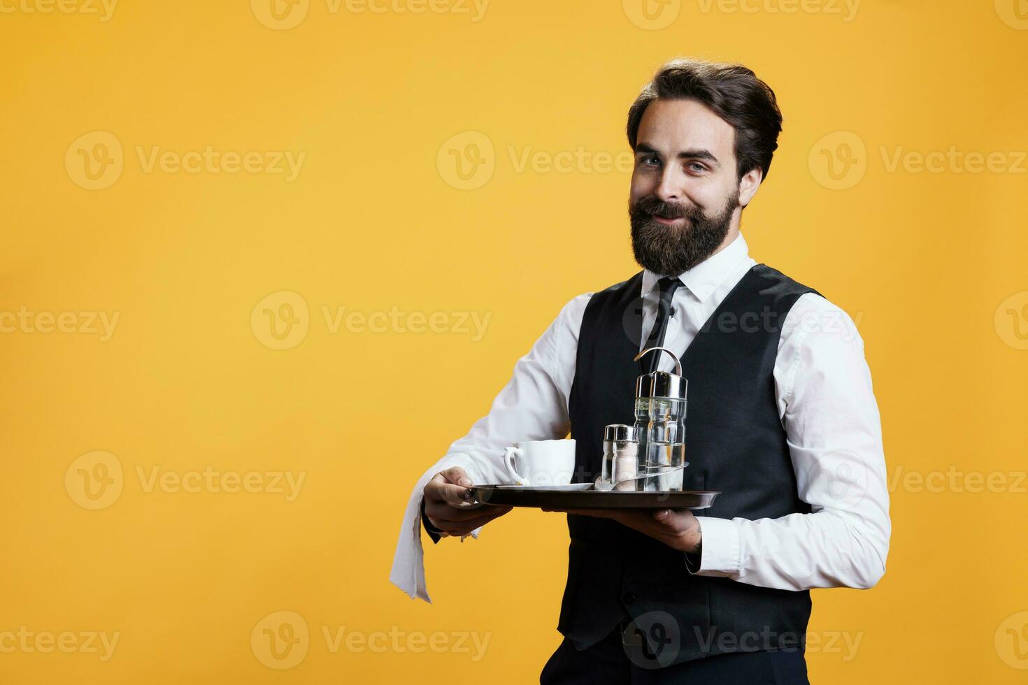 Elegant man butler holding tray to put salt and pepper on table at restaurant, carrying cup of coffee on platter for customers in studio. Professional luxurious waiter posing on camera. photo