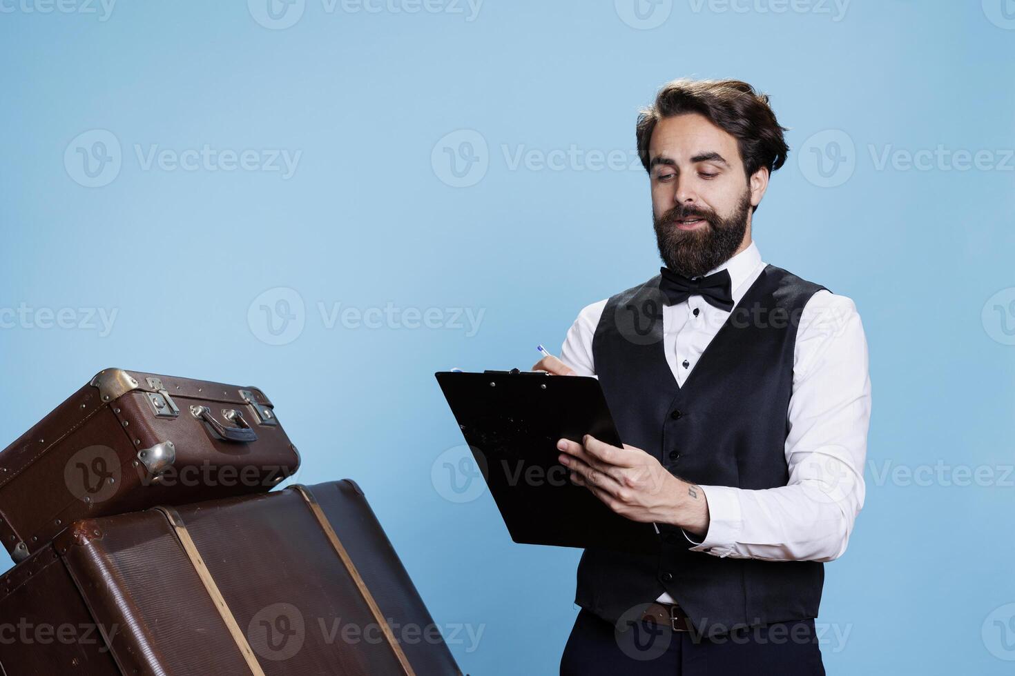 Bellboy checking guest list in studio, reading professional hotel record papers on clipboard. Young person works as doorman verifying register file or log while he stands against blue background. photo