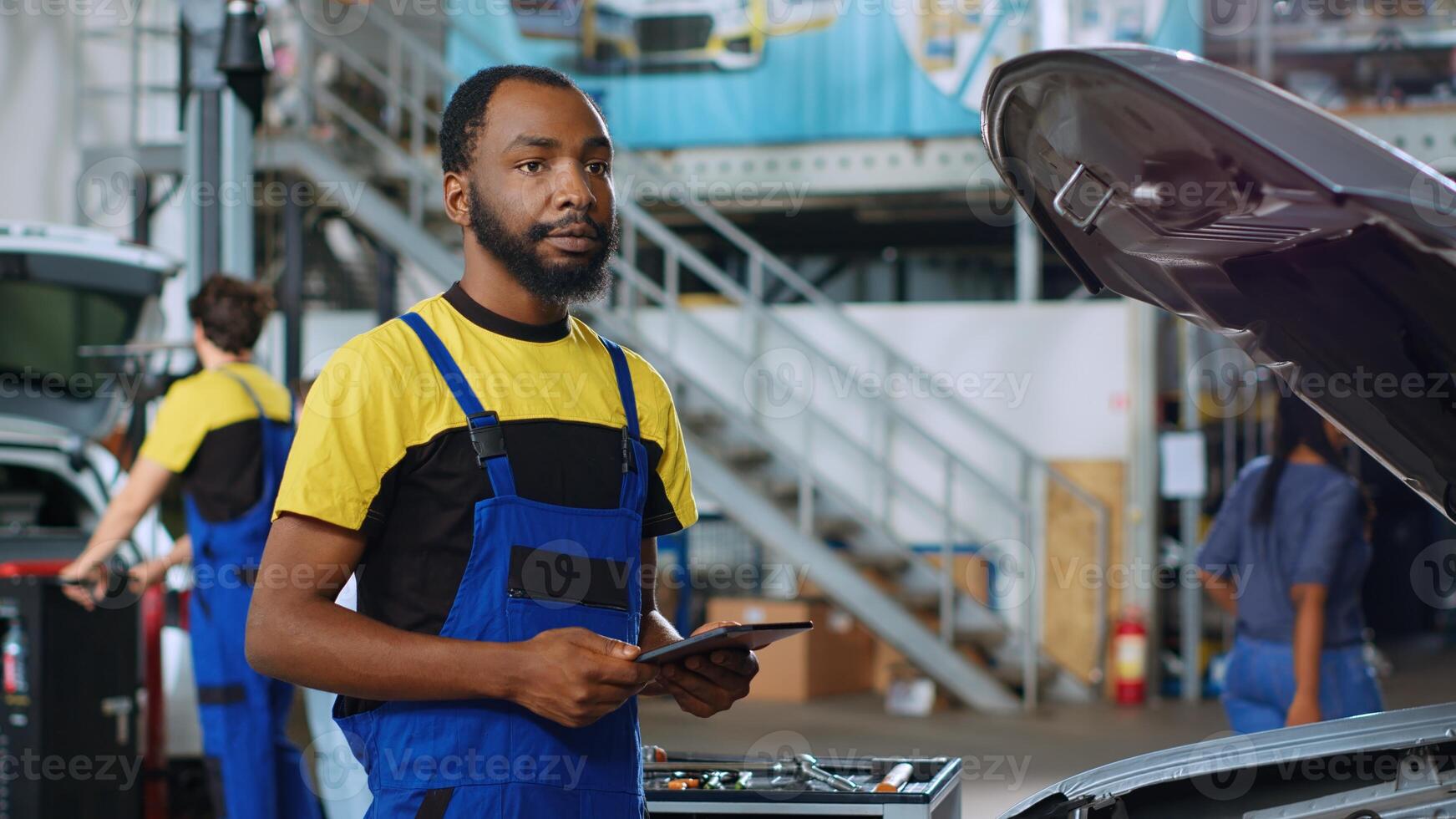 Portrait of happy engineer in car service ordering new parts for damaged vehicle using tablet. African american expert looking online for components to replace old ones in defective automobile photo