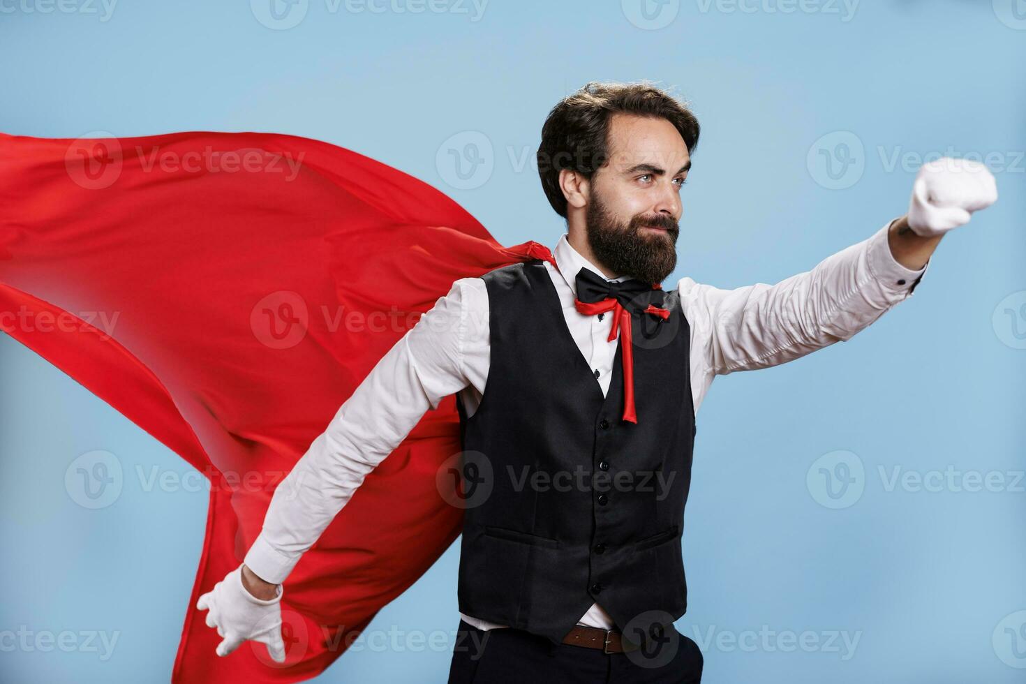 Determined adult posing as superhero while he wears red mantle against blue background in studio, professional person acting like a superhuman. Man with formal attire and cartoon cape. photo