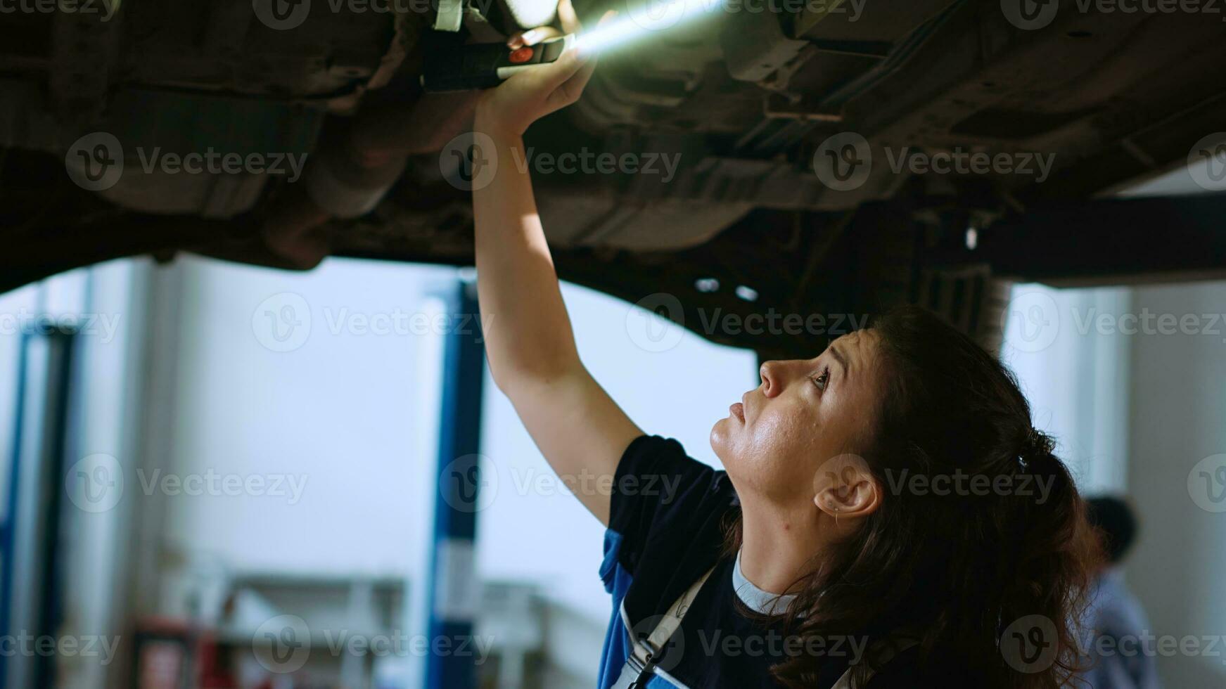 Woman working on suspended car in garage, checking parts during annual checkup. Professional in repair shop workplace walking underneath vehicle, scanning it using work light photo