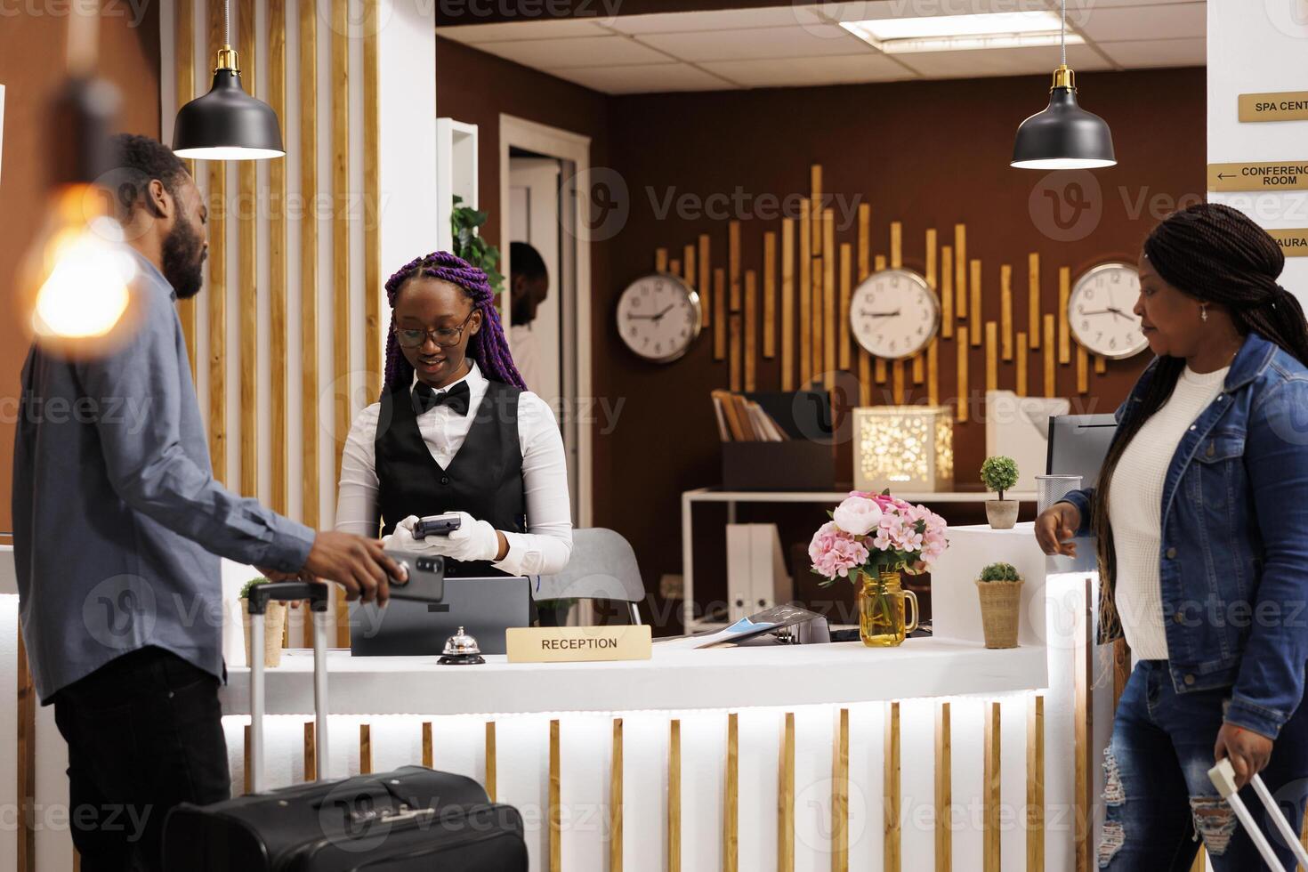 Young African American couple standing at reception counter paying for hotel room while checking out. Tourists using mobile phone to pay, making nfc payment while registering at front desk photo