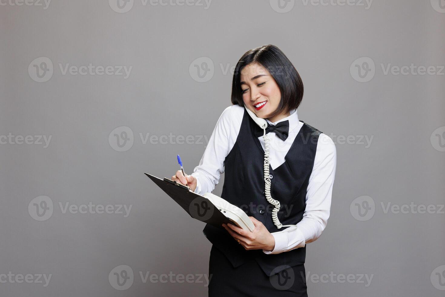 Smiling asian waitress listening to manager instructions on landline phone and taking notes in clipboard. Catering service woman worker speaking on telephone and writing checklist photo