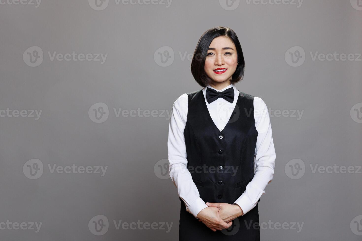 Smiling attractive asian waitress wearing uniform with bow tie standing and looking at camera. Restaurant professional receptionist posing for studio portrait on gray background photo