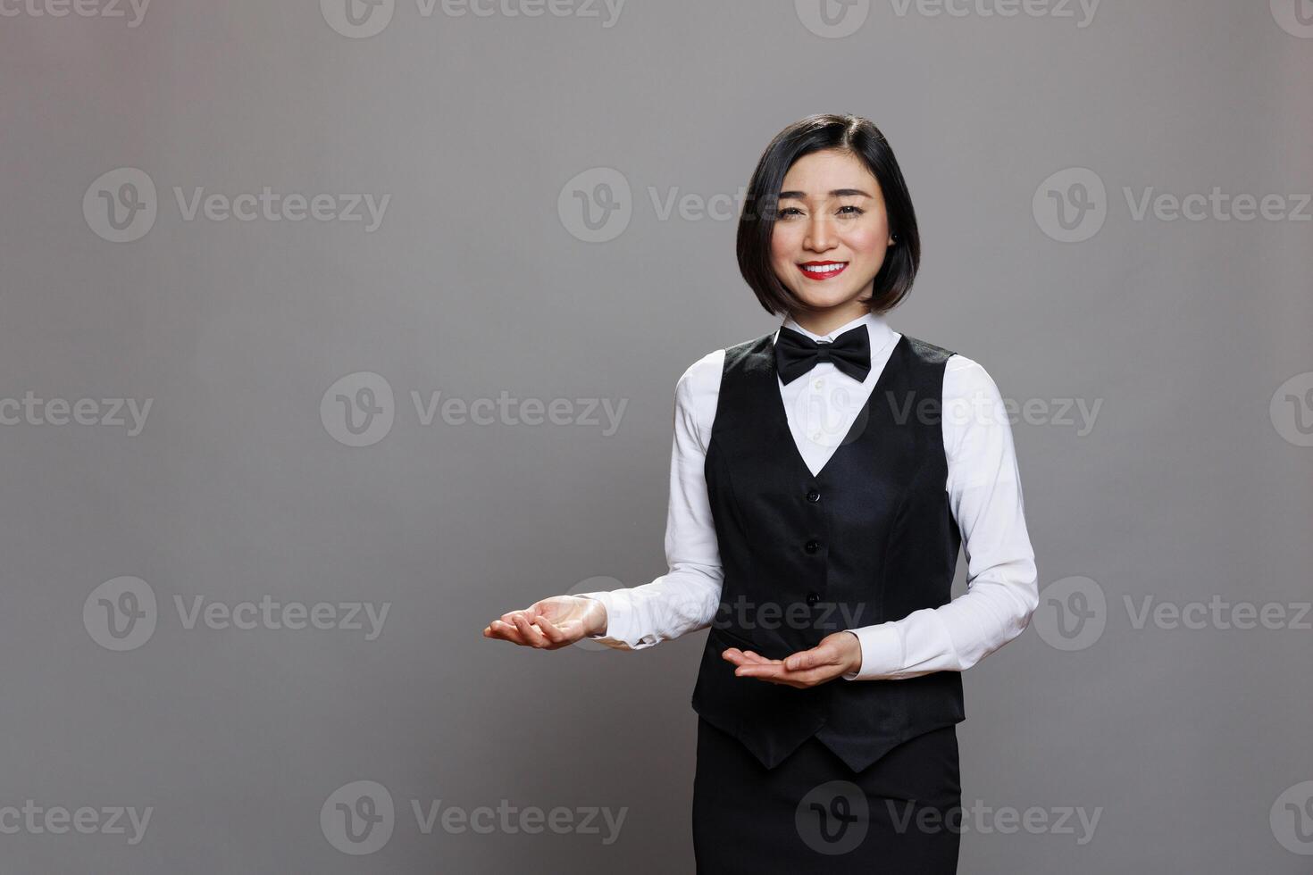 Smiling asian woman waitress wearing restaurant uniform showing with hands and greeting guests portrait. Professional receptionists welcoming customers and looking at camera photo