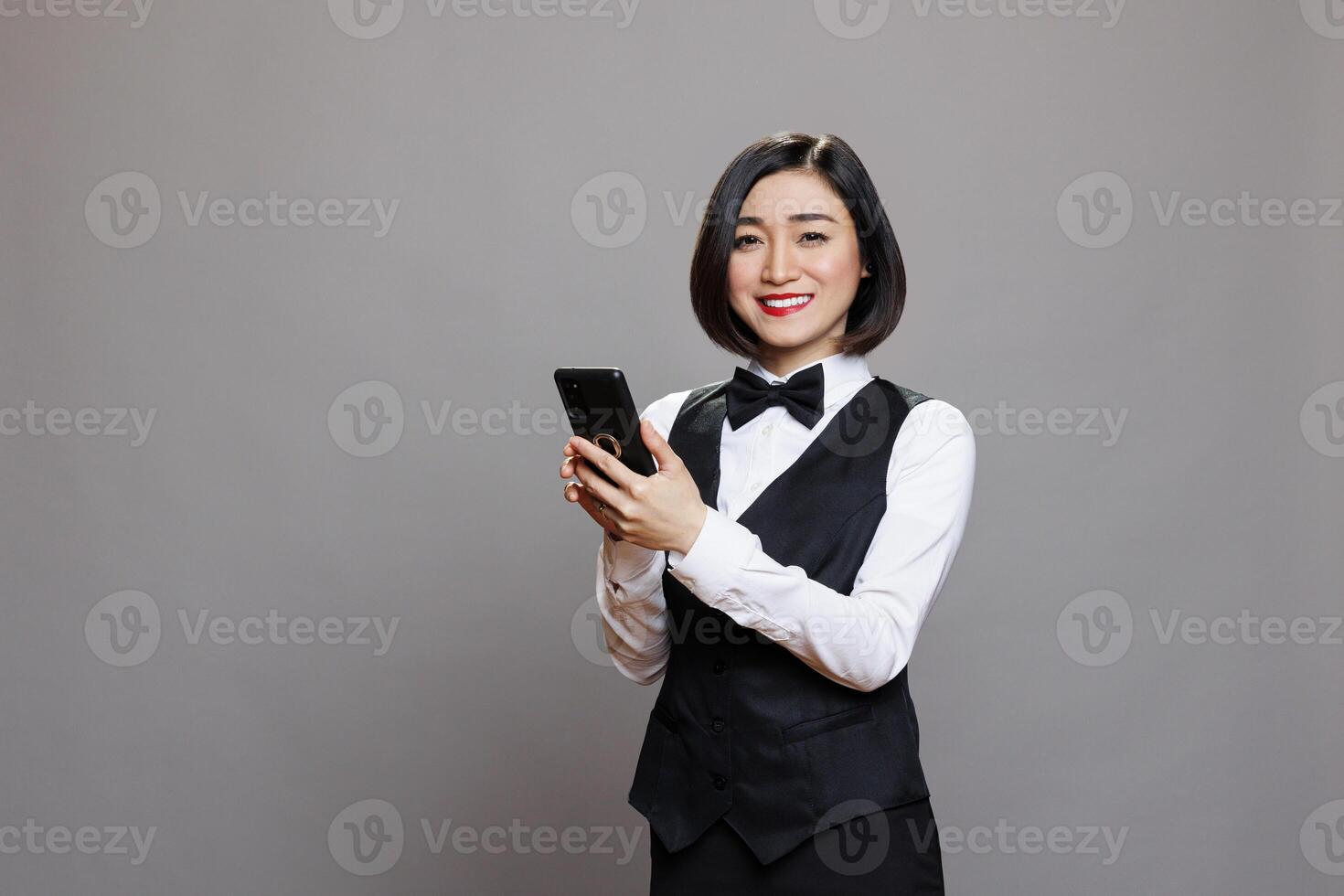 Smiling asian waitress using smartphone and looking at camera with friendly facial expression. Cheerful restaurant receptionist in professional uniform holding mobile phone portrait photo