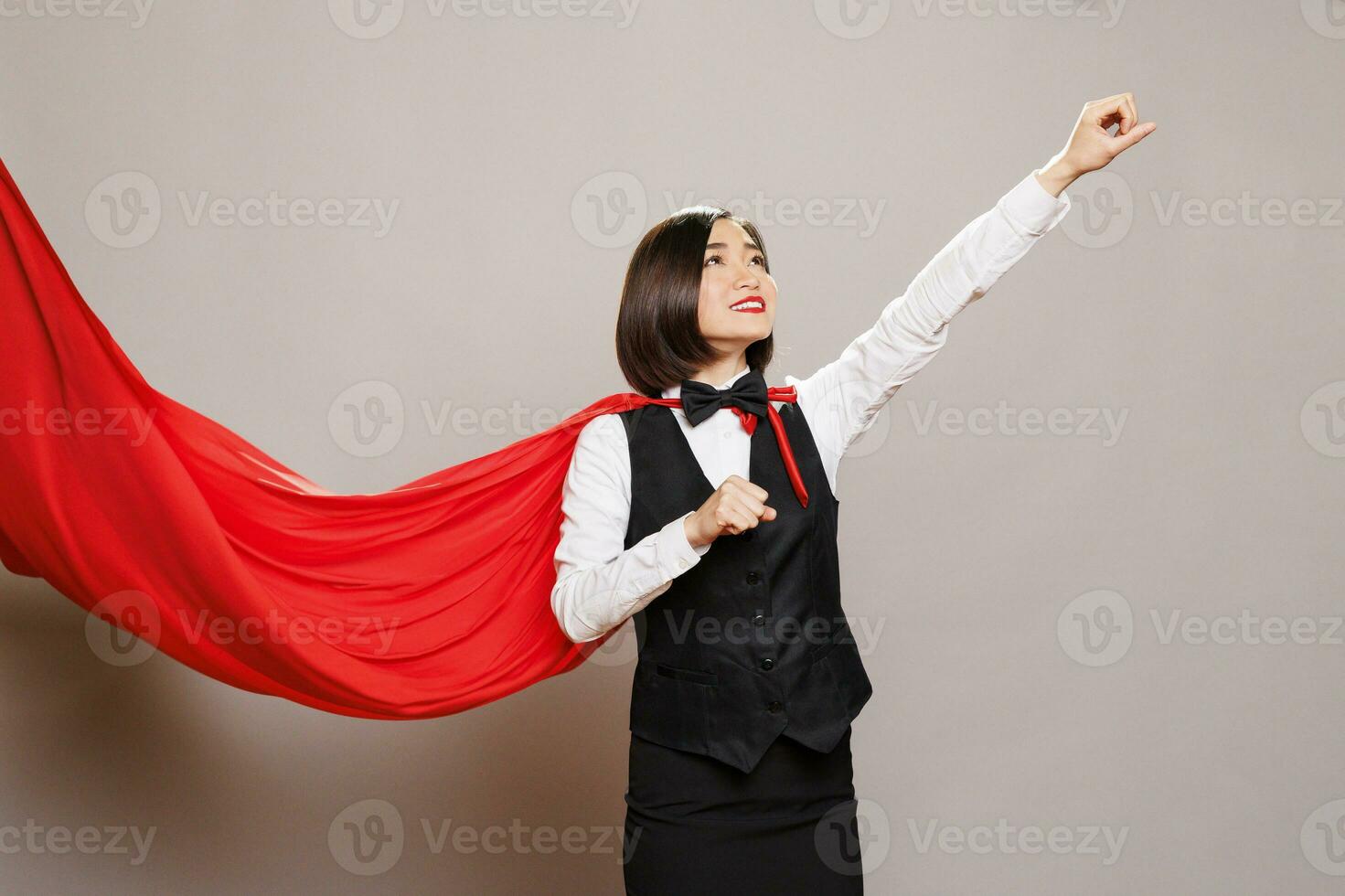 Asian waitress wearing superwoman cloak raising clenched fist and looking upwards. Catering service woman employee flying in fluttering hero red cape while posing in studio photo